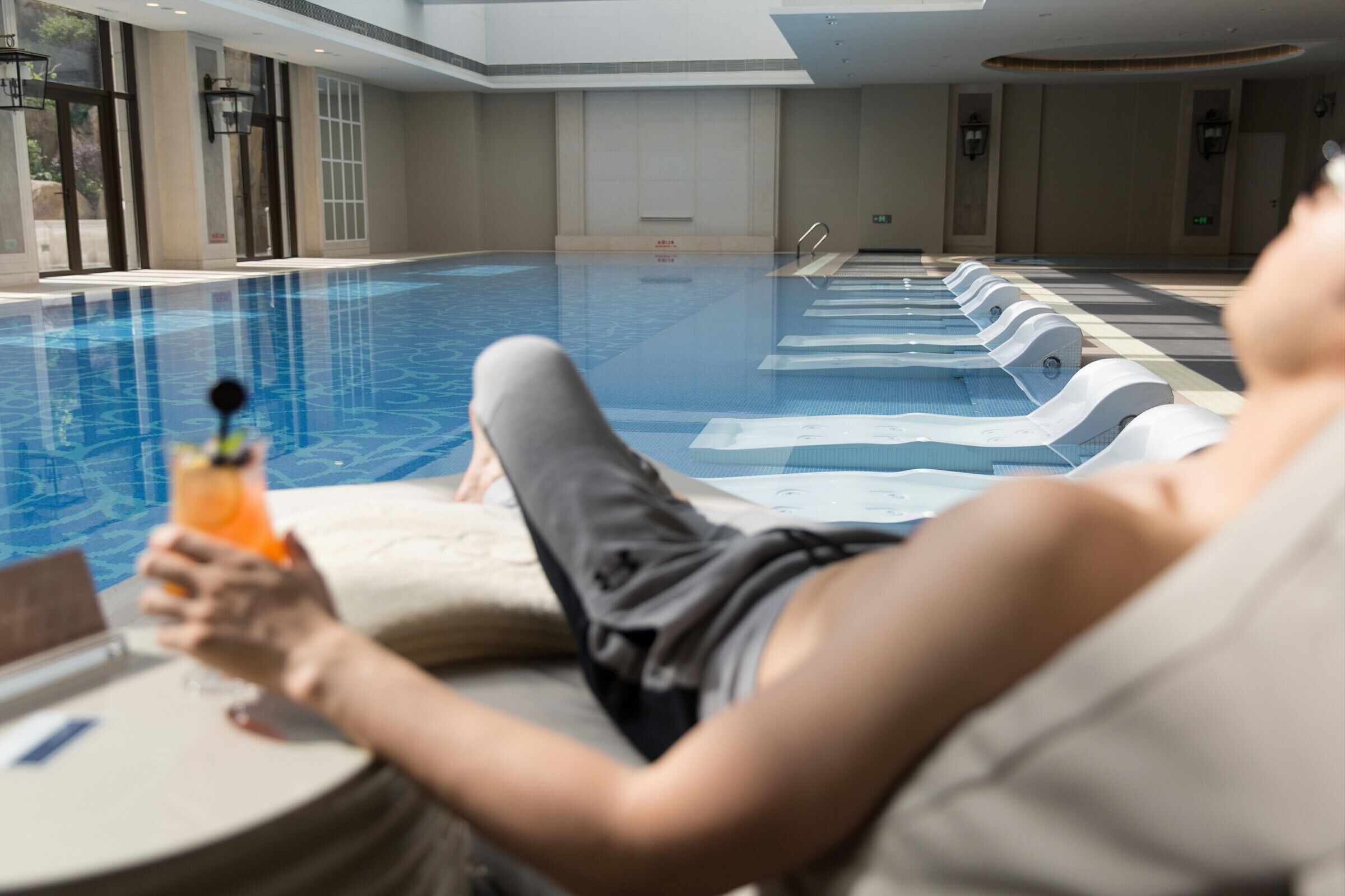 Una persona descansando junto a una piscina con sillones reclinables, agua cristalina y un diseño interior moderno.