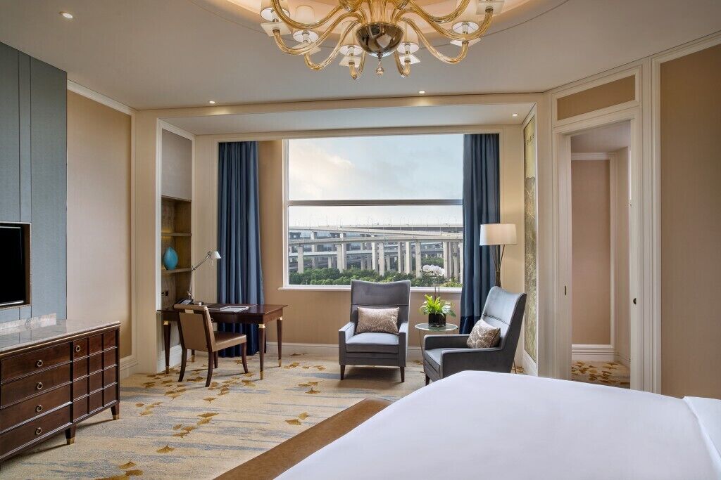 A hotel room with a large bed, a desk, two armchairs, a television, and a view of a cityscape through a large window.