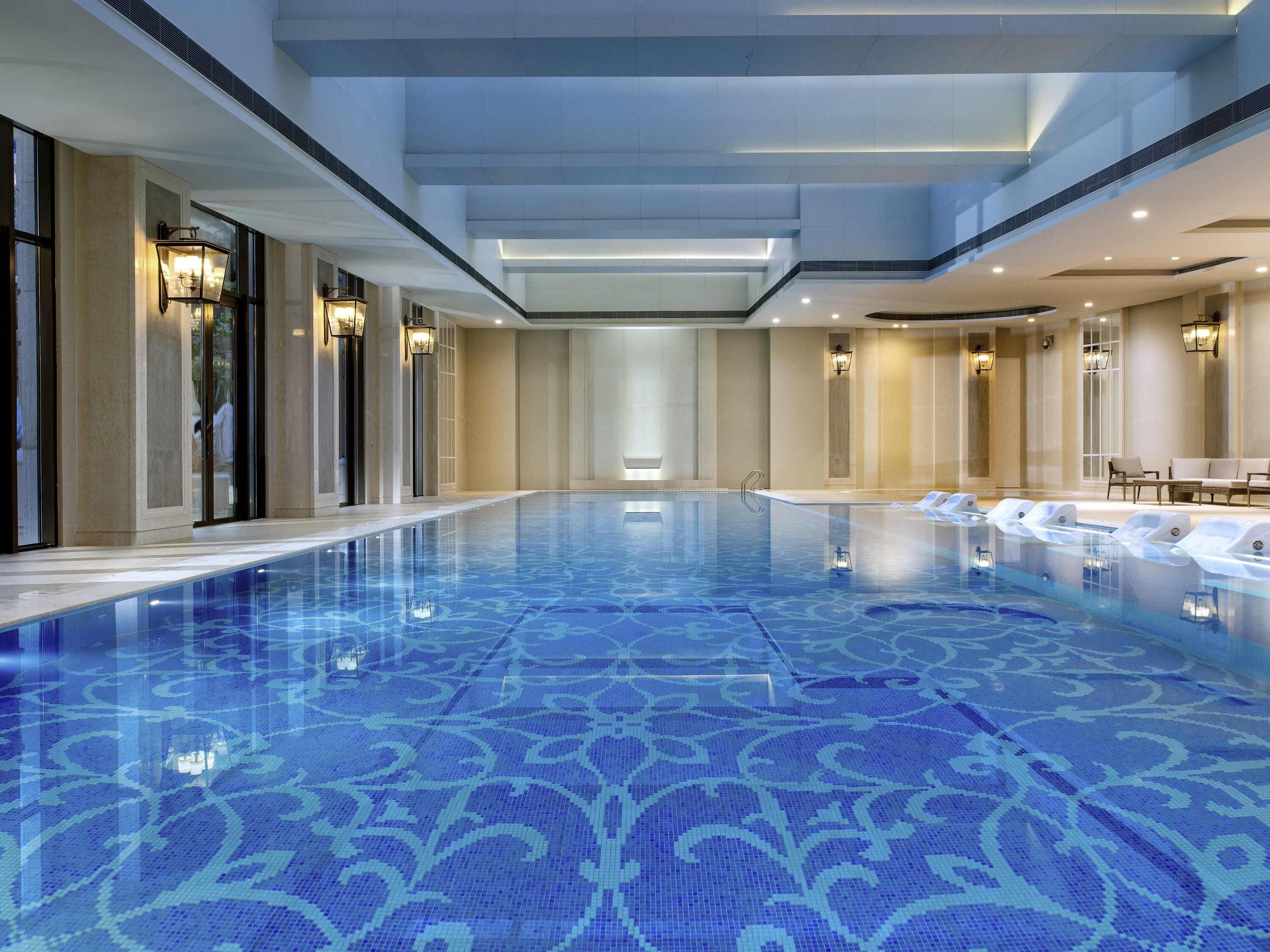 A large indoor swimming pool with a blue patterned floor, surrounded by walls with light fixtures and seating areas.
