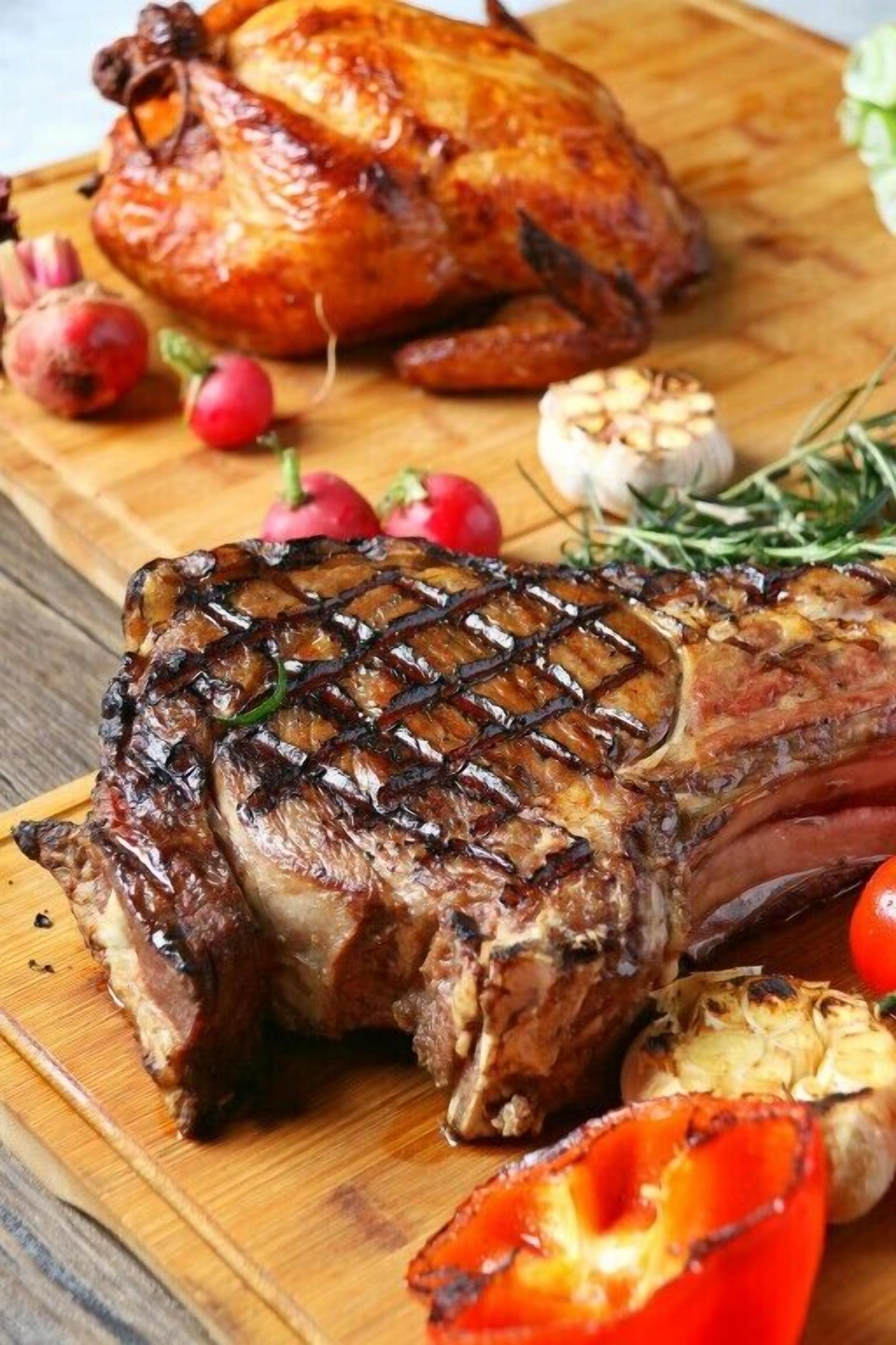 Gebratenes Hähnchen und Steak auf einem Holzbrett mit Knoblauch, Radieschen und einer Tomate.