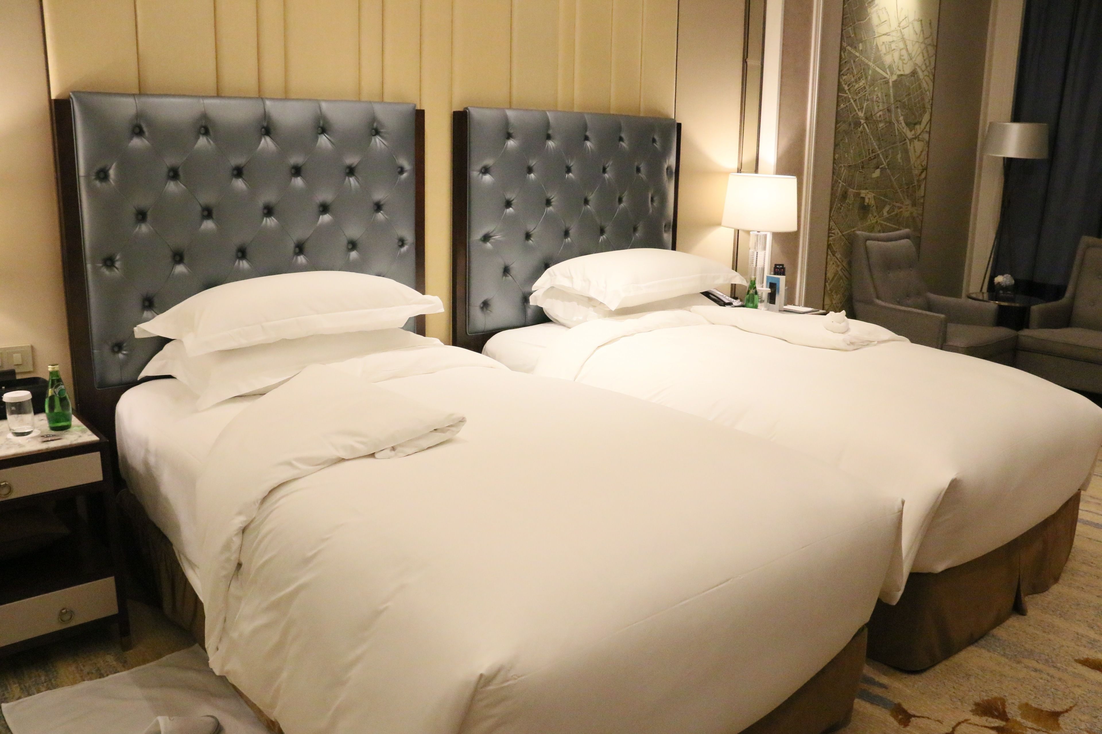A hotel room with two beds, each with a tufted headboard, white bedding, and a nightstand with a lamp and a bottle.