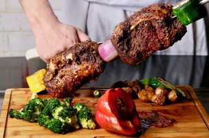 A person grilling meat skewers on a wooden cutting board with vegetables.