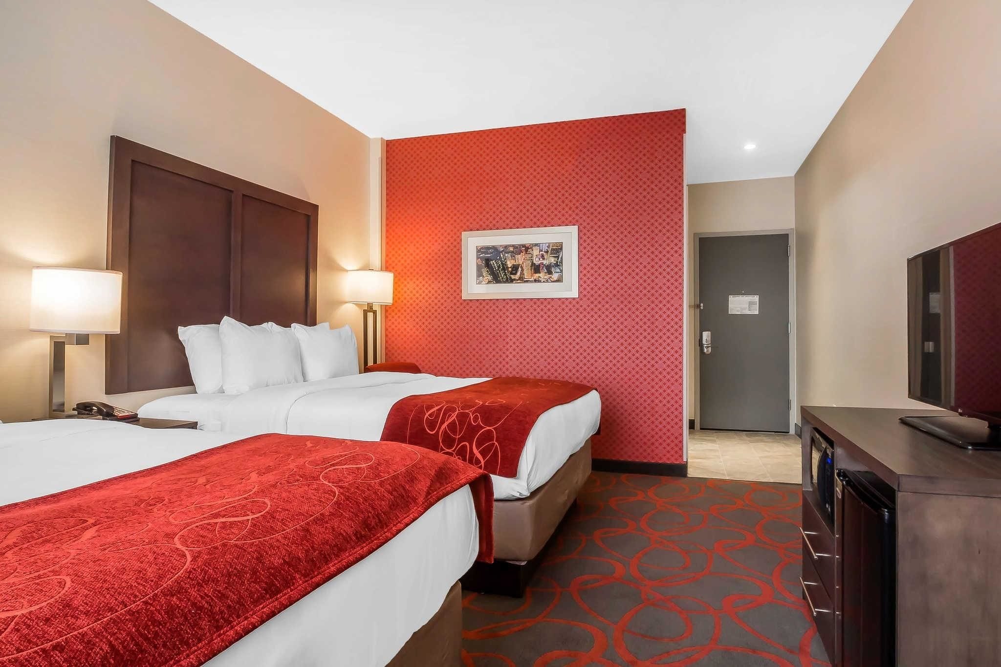 Une chambre d’hôtel avec deux lits, un mur décoré d’un motif rouge, un tableau encadré, un bureau avec une télévision et une porte.