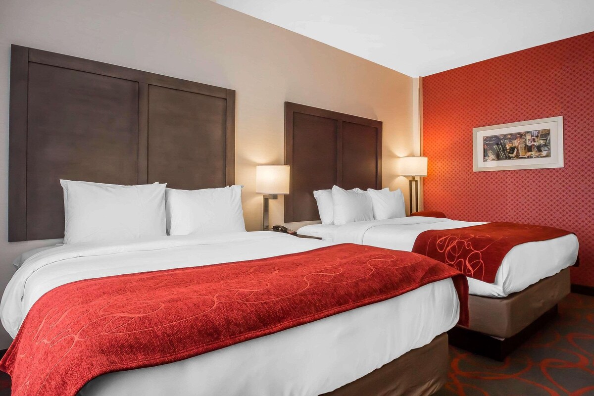 A hotel room with two beds, each with white bedding and a red blanket. There are two bedside tables with lamps, and a framed picture on the wall.