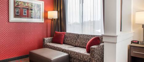 A hotel room with a patterned sofa, a small ottoman, a framed picture on the wall, and a window with curtains.