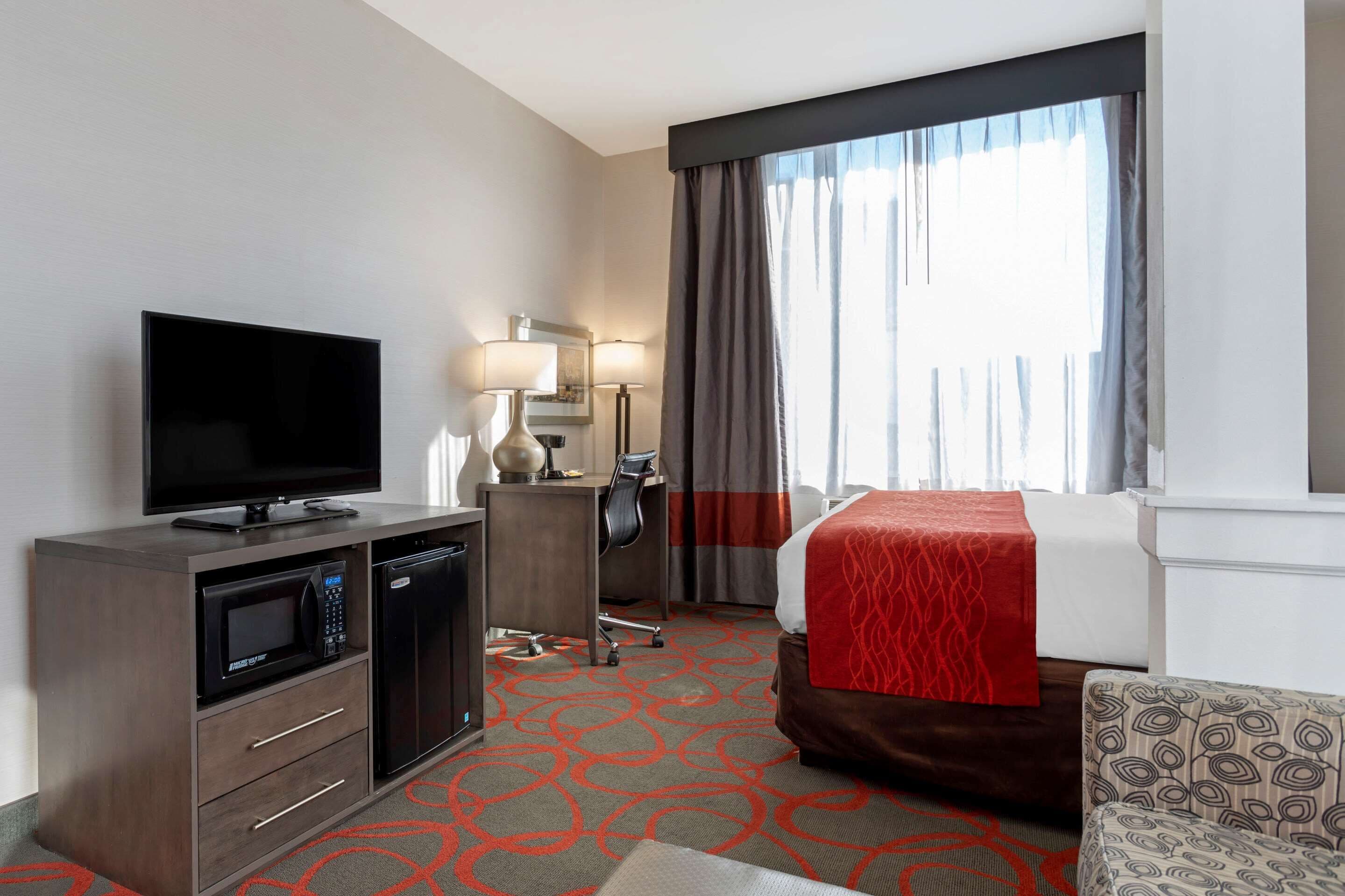 A hotel room with a flat-screen TV, a microwave, a desk with a chair, a bed with a red blanket, and a patterned armchair.
