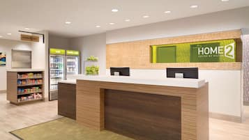 A reception area with a wooden reception desk, a "HOME 2" sign, a refrigerator, and a snack shelf.