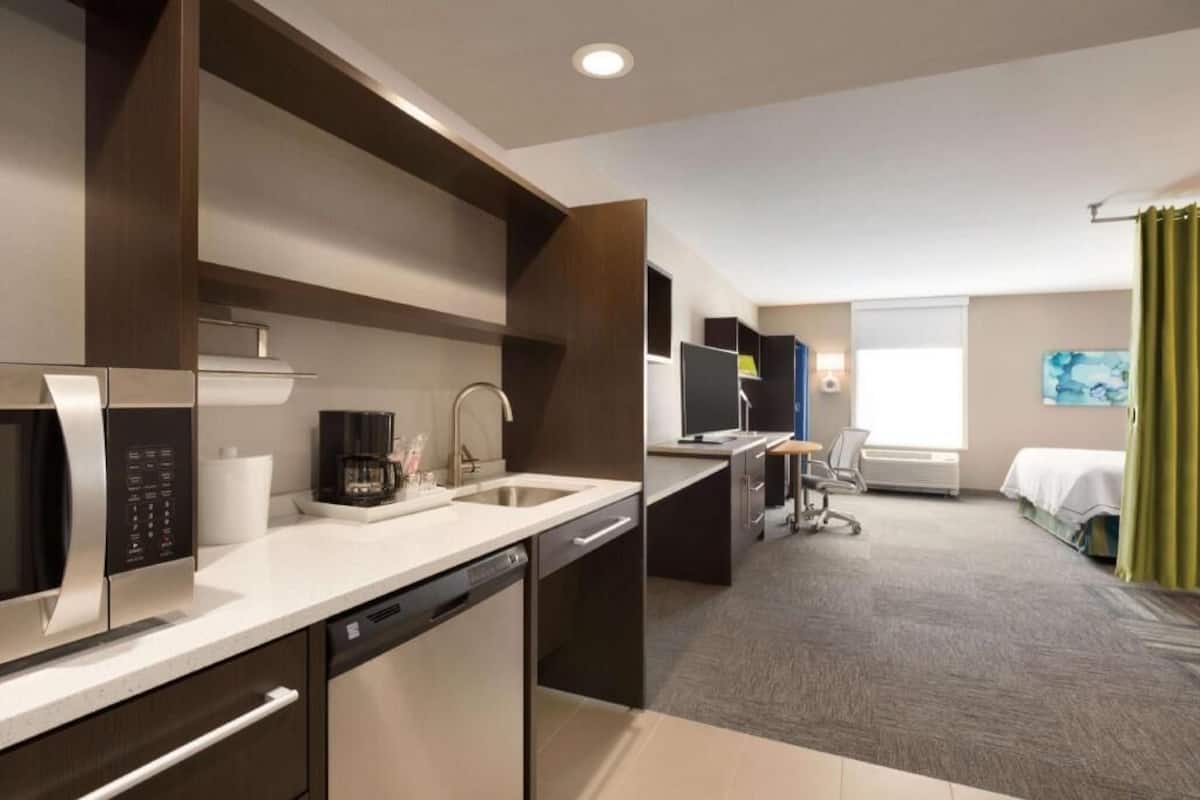A modern hotel room with a kitchenette, a bed, a desk, and a window with curtains.