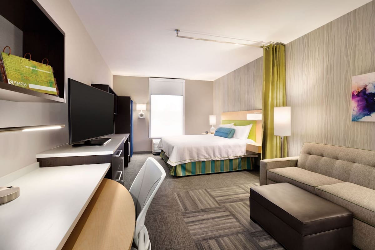 A modern hotel room with a bed, a sofa, a desk, a TV, and a bathroom.