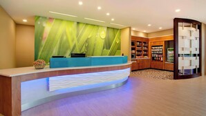 Un área de recepción de diseño moderno, con mostrador de recepción, una pared con diseño de hojas verdes y una sección con estanterías que contienen diversos objetos.