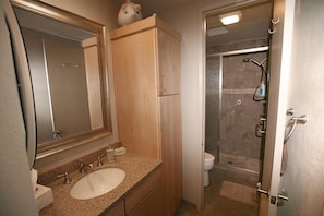 A bathroom with a sink, mirror, wooden cabinet, and a shower area. - Sunset Suite (Oceanside)