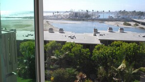 Vista desde una ventana sobre una playa, un puerto deportivo y un estacionamiento con árboles y vegetación al frente.