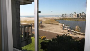 A view from a balcony overlooking a beach, a body of water, and a distant shoreline with buildings and palm trees. - Harbor Cottage (Oceanside)