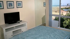A bedroom with a bed, a TV on a white cabinet, a large window with a beach view, and three framed pictures on the wall. - Harbor Cottage (Oceanside)