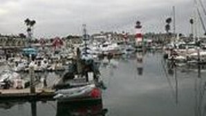 A marina with multiple boats docked, a prominent lighthouse, and a cloudy sky. - Harbor Cottage (Oceanside)
