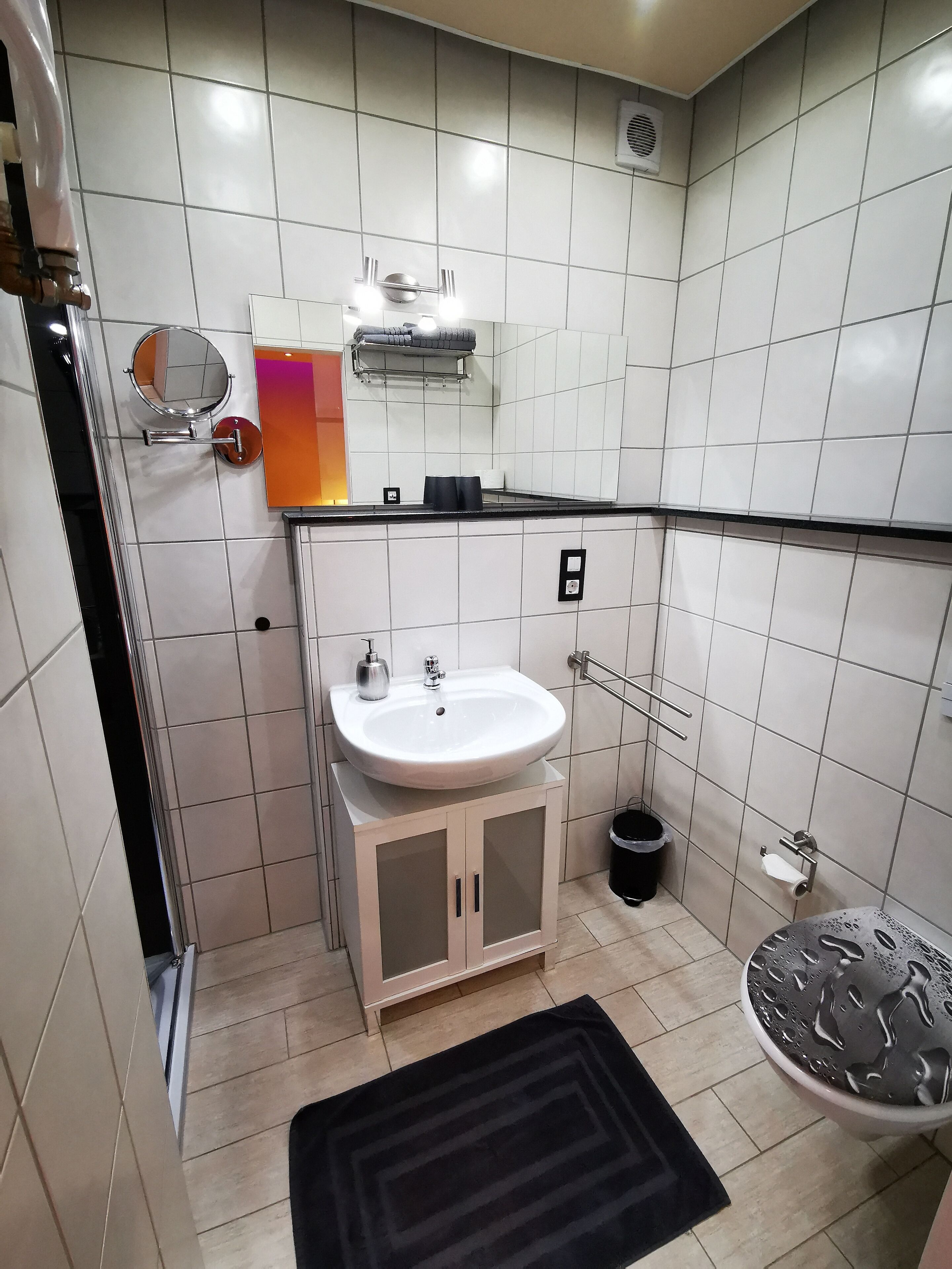 A bathroom with white tiled walls, a sink with a mirror, a towel rack, a black trash bin, and a black rug.