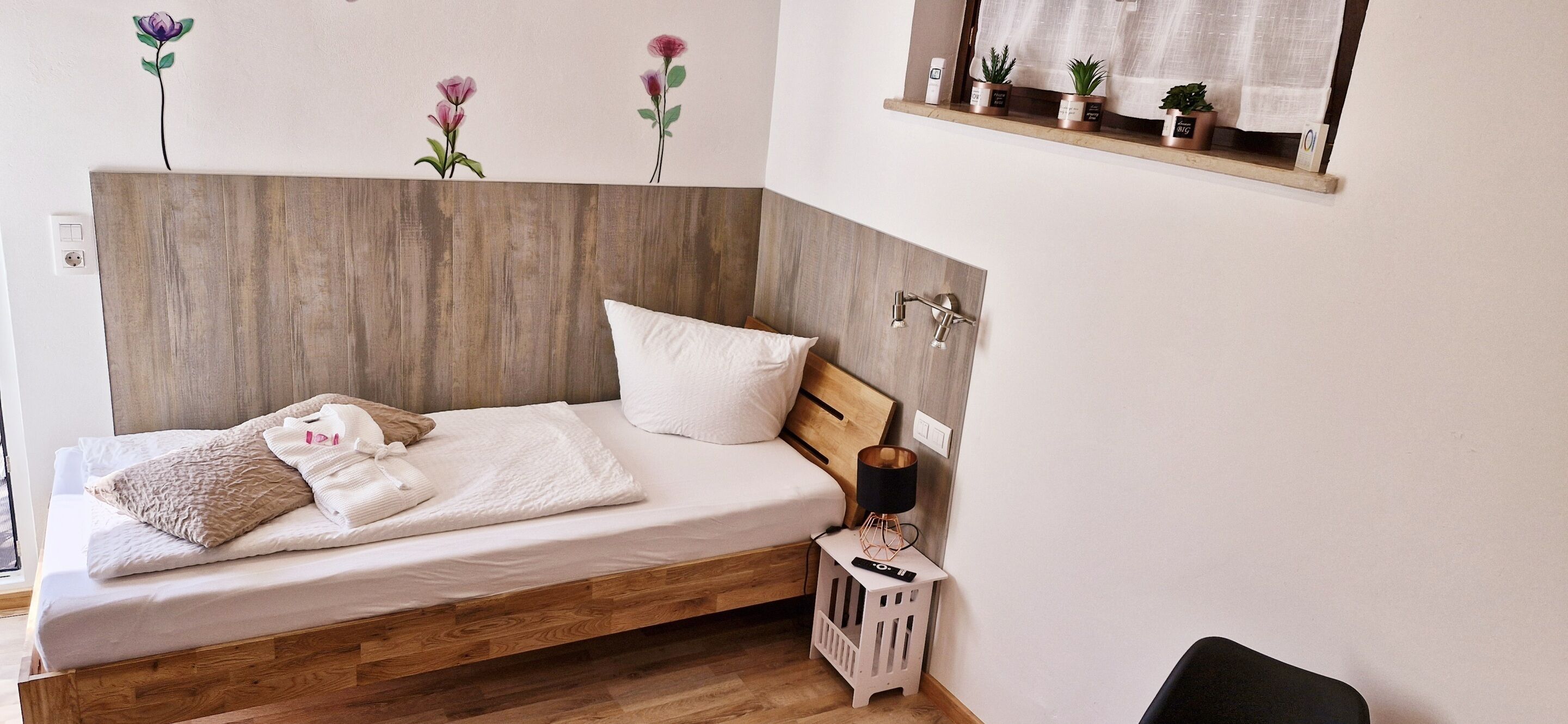 A bedroom with a wooden headboard, a bed with white bedding, a bedside table with a lamp, and a window with a view of the outside.
