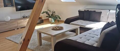 A modern loft-style living room with a slanted ceiling, a large flat-screen TV, a wooden coffee table, a sofa, and a potted plant.