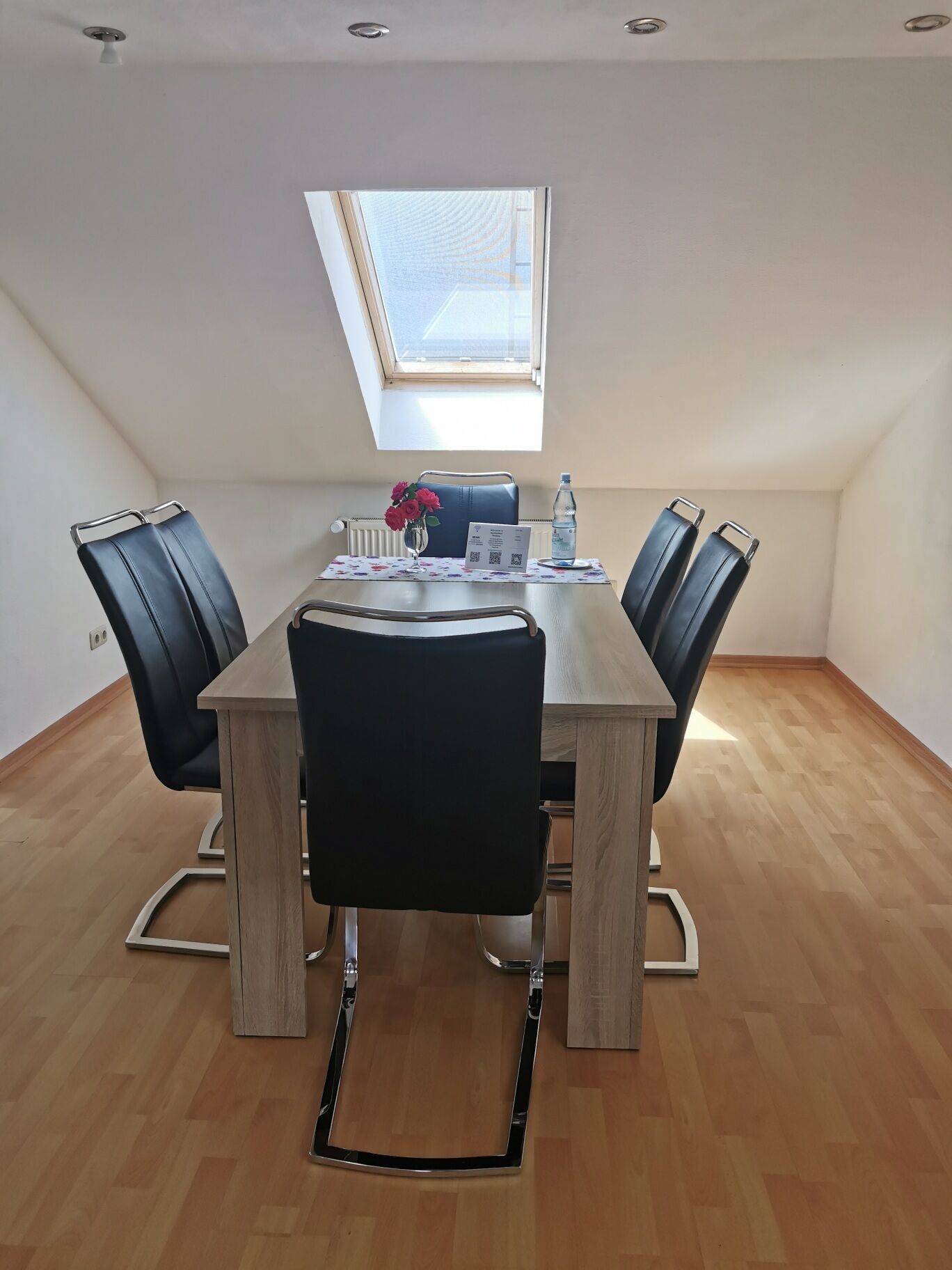 A modern conference room with a wooden table, black chairs, a skylight, and a vase with flowers.