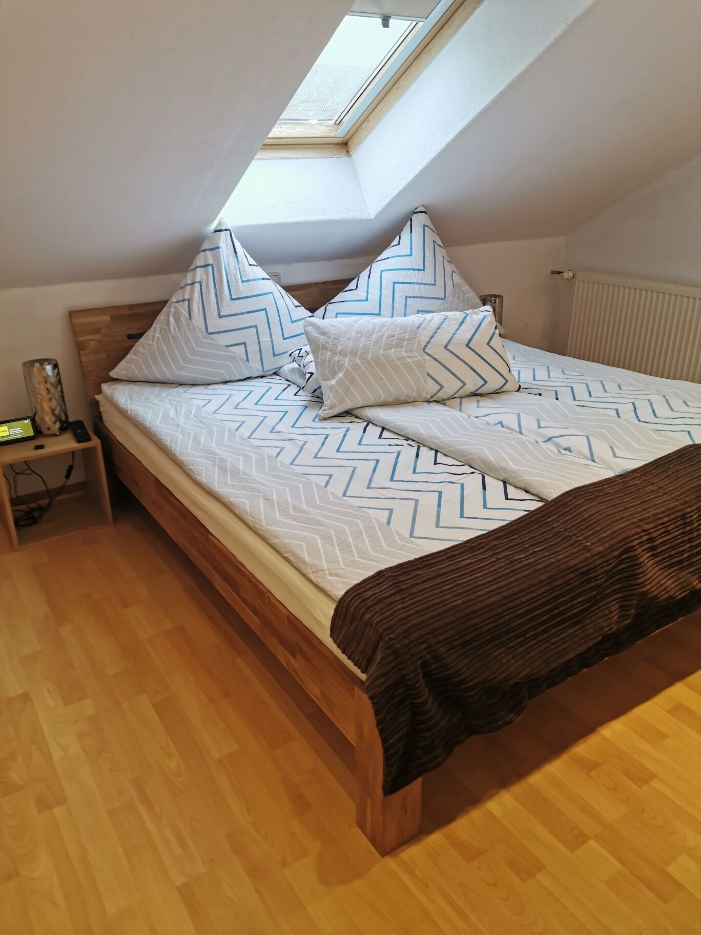 A bedroom with a skylight, a wooden bed frame, a nightstand, and a bedside table.