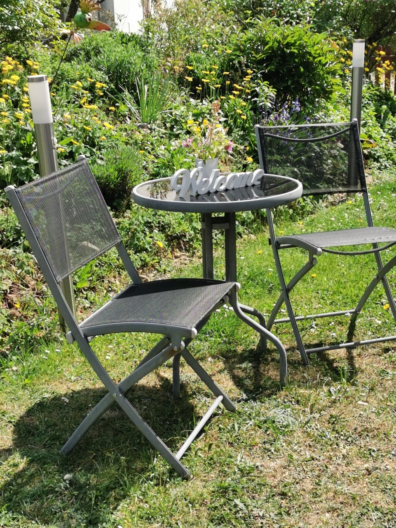 An outdoor seating area with a round table and two chairs, surrounded by greenery and flowers.