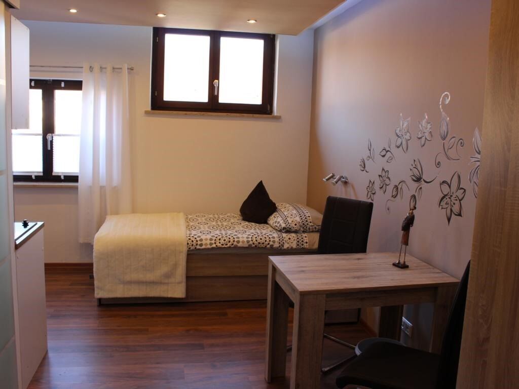 Een compacte kamer met een bed, een klein tafeltje en een stoel. Er is een raam met gordijnen, een wandlamp en een decoratief wandobject.