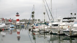 A marina with multiple boats docked, a lighthouse, and a building with palm trees in the background. - Steps to the Sand (Oceanside)