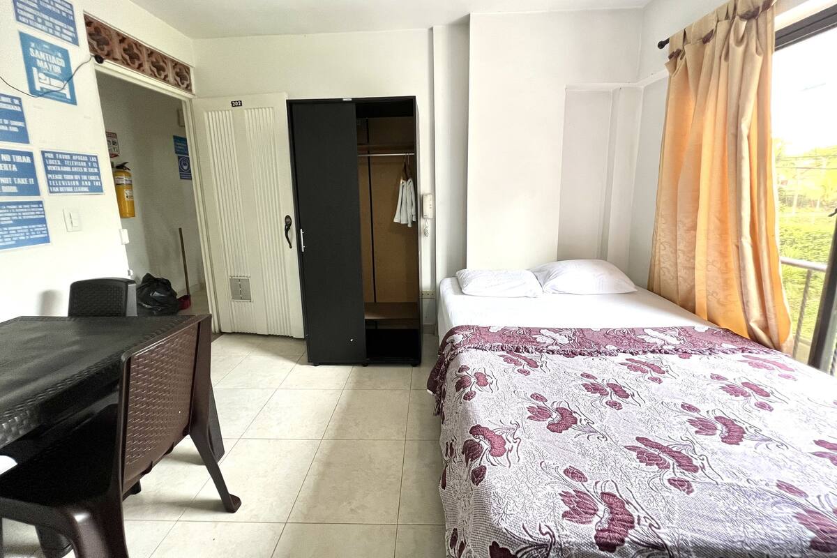Hotellihuoneessa on sänky, työpöytä ja vaatekaappi. Seinällä on kylttejä ja ikkunassa on verhot.