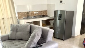 A living room with a grey sofa, a remote control on it, a kitchen area with a fridge, and a tiled floor. - Casa Santiago Mayor (Cali)