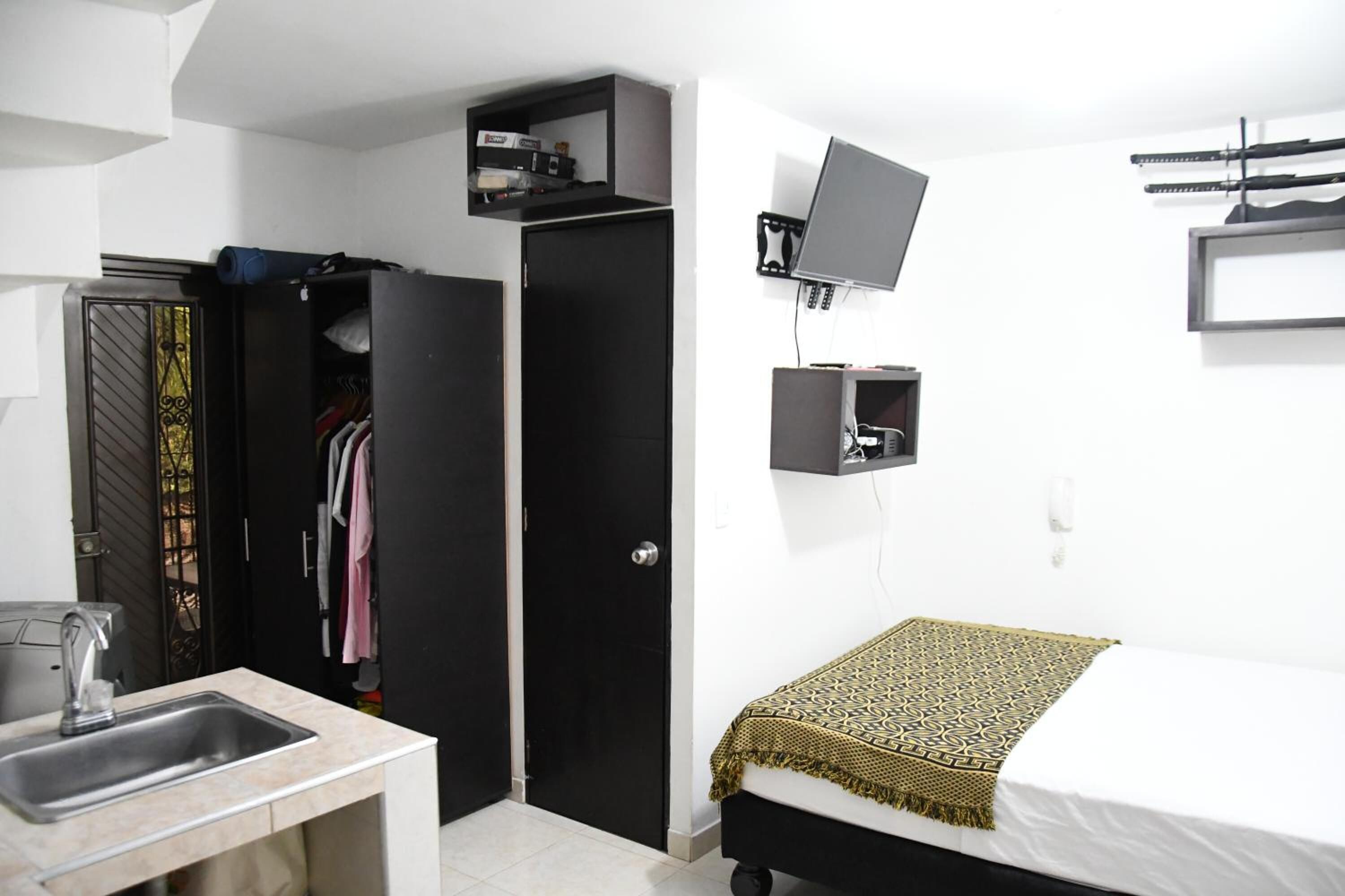 A compact room with a bed, a desk, a chair, a wardrobe, and a TV mounted on the wall.