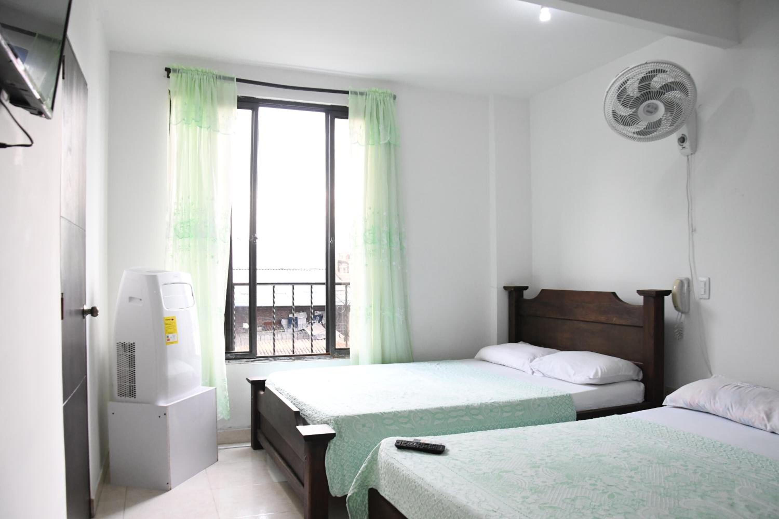 Habitació amb dos llits, una finestra amb cortines, un radiador portàtil i un ventilador fixat a la paret.