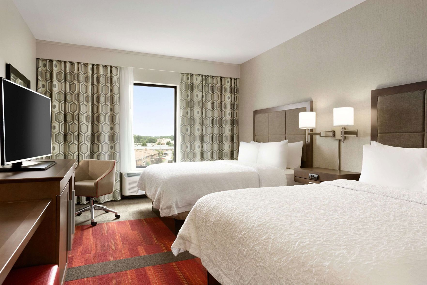 A hotel room with two beds, a desk with a TV, a chair, and a window with curtains.