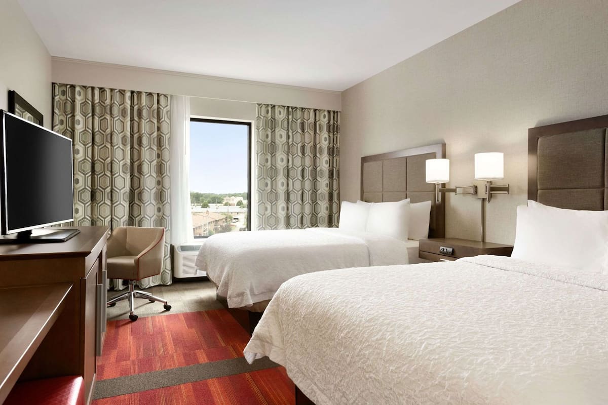 A hotel room with two beds, a desk with a TV, a chair, and a window with curtains.