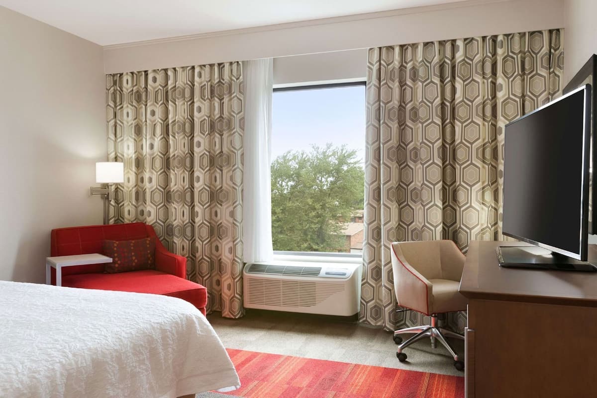 Une chambre d’hôtel avec un lit, un canapé rouge, un bureau avec une télévision et une fenêtre ornée de rideaux à motifs.