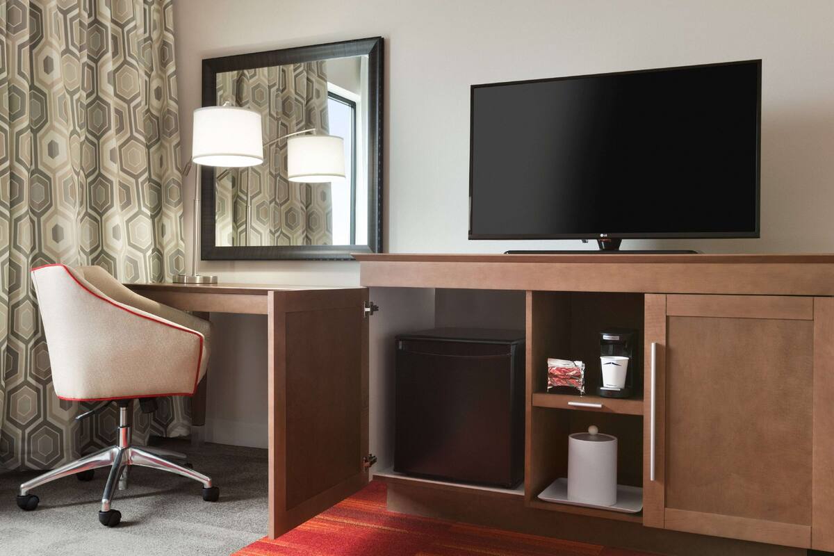Une chambre d’hôtel équipée d’un bureau en bois, d’une télévision, d’une chaise, d’un miroir et d’un petit réfrigérateur.