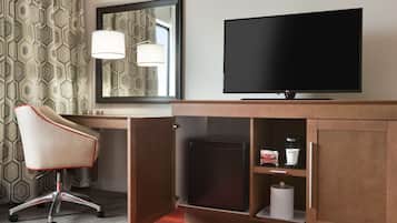 A hotel room with a wooden desk, a television, a chair, a mirror, and a small refrigerator.