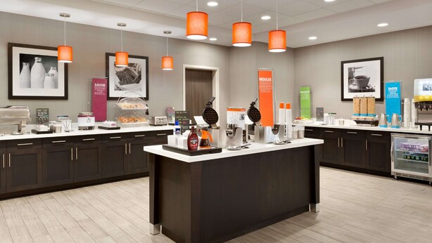 A modern kitchen area with a central island, overhead orange pendant lights, and various kitchen appliances and food items. - Hampton Inn North Olmsted Cleveland Airport (North Olmsted)