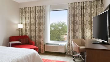 A hotel room with a bed, a red sofa, a desk with a TV, a chair, and a window with patterned curtains.