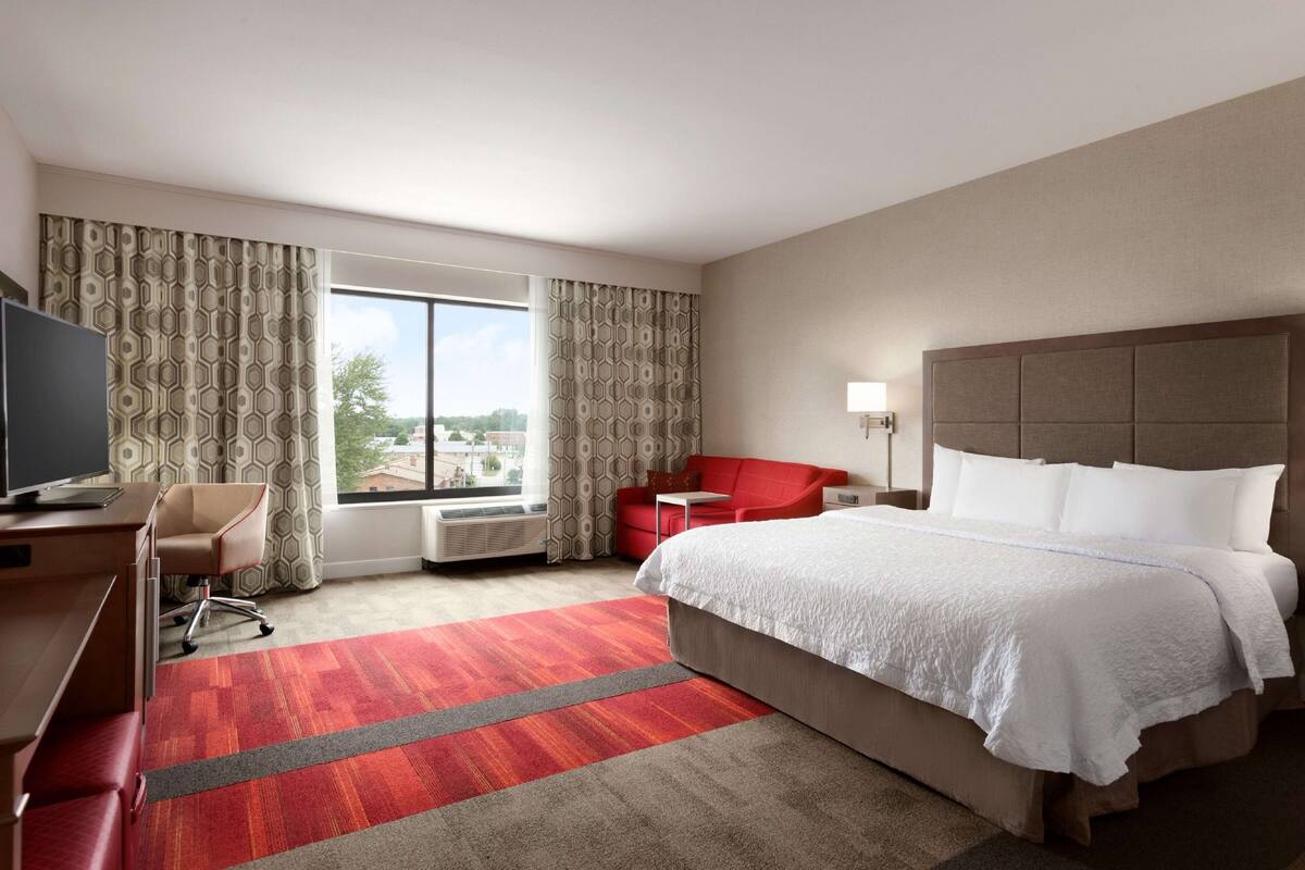 Une chambre d’hôtel avec un grand lit, un bureau avec une chaise, un canapé rouge et une fenêtre avec des rideaux.