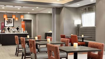 A hotel dining area with multiple tables and chairs, a counter with amenities, pendant lighting, and a large window with curtains.
