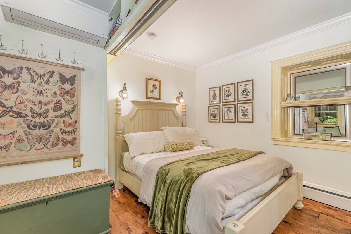 Un dormitorio con cama, lámpara de pared, cuadro con mariposas y ventana con vista a follaje.