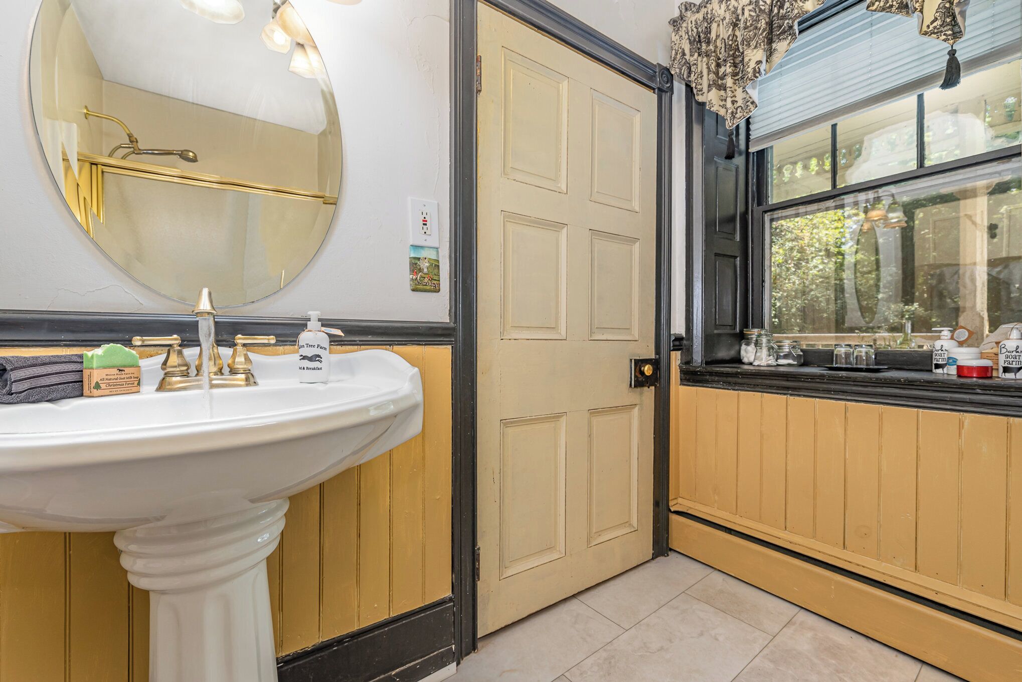 Une salle de bain avec un lavabo sur colonne, un miroir rond, un porte-serviettes, une porte avec une fenêtre et un distributeur de serviettes.