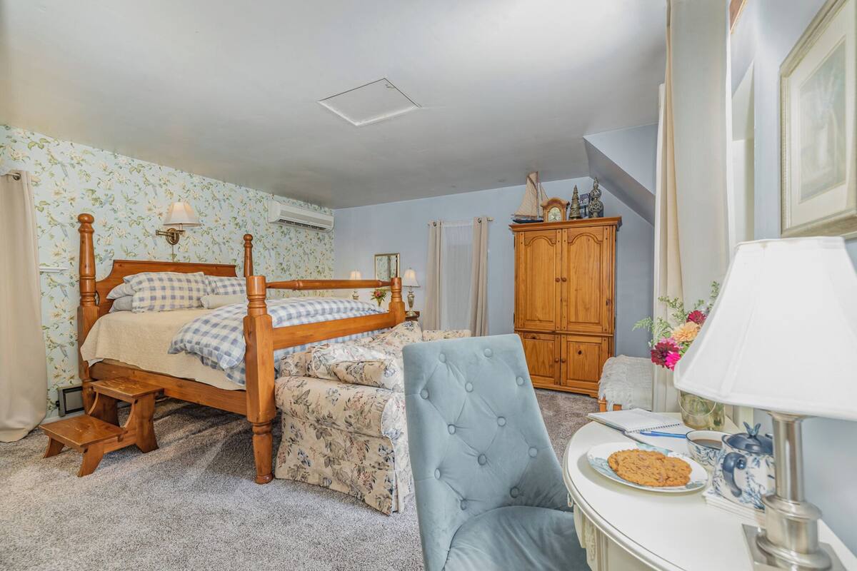 Un dormitorio con una cama de madera, una silla, una mesita con un plato y una lámpara, y un armario.