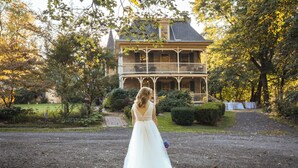 Une mariée, vêtue d’une robe blanche, se tient devant une grande maison historique dotée d’un balcon.