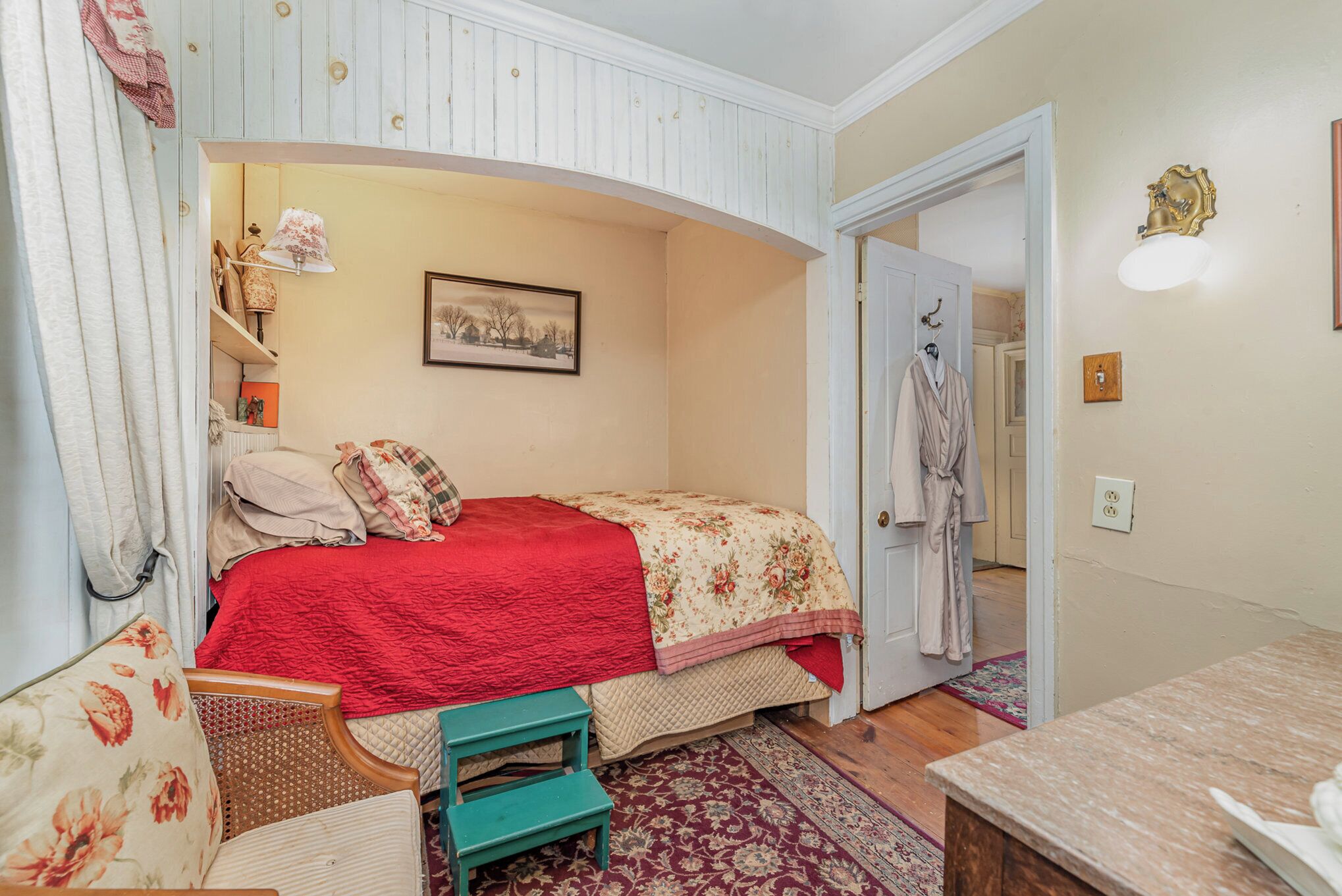A bedroom with a bed, a chair, a bench, a lamp, a picture on the wall, and a door leading to another room.
