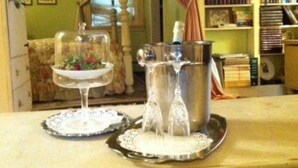 Une table avec une carafe et un verre d’eau, un bol de nourriture et un objet décoratif, dans une pièce où se trouvent une bibliothèque et une fenêtre.