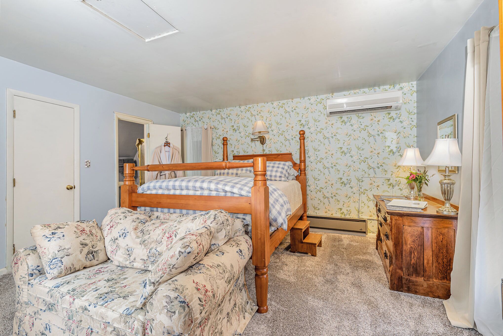 A bedroom with a wooden four-poster bed, a floral sofa, a wooden dresser, and a nightstand with a lamp.