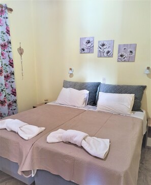 A double bed with a beige bedspread and two pillows, a nightstand with a lamp, and floral curtains. - Amadeus Hawaii Studios Apts (Corfu)
