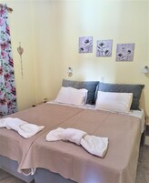 Una cama doble con colcha color beige y dos almohadas, una mesita de noche con lámpara y cortinas con estampado floral.