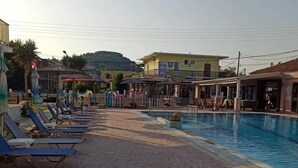 A swimming pool area with lounge chairs and umbrellas, a restaurant, and a hill in the background. - Amadeus Hawaii Studios Apts (Corfu)